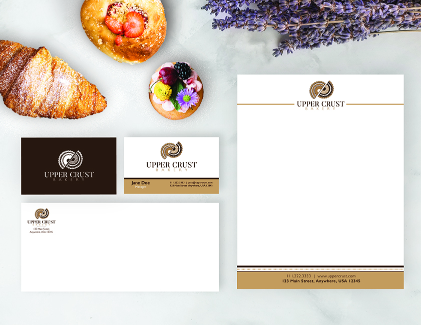 Flat-lay image of Upper Crust Bakery letterhead, envelope, and business card design, each featuring the custom business logo, on a white marble background near delicious pastries and a bouquet of lavender.